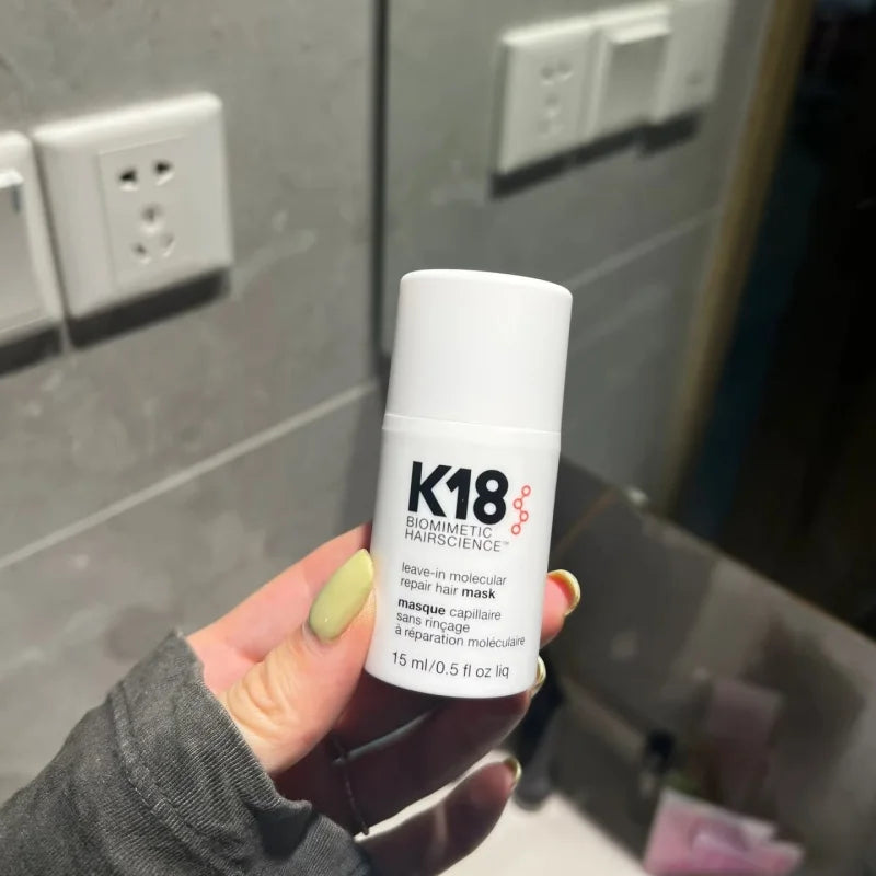 Hand holding K18 Leave-In Molecular Repair Hair Mask 15ml mini bottle in bathroom setting