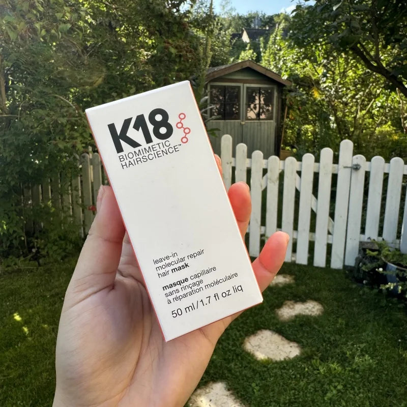 Hand holding K18 Leave-In Molecular Repair Hair Mask 50ml product box in outdoor garden setting